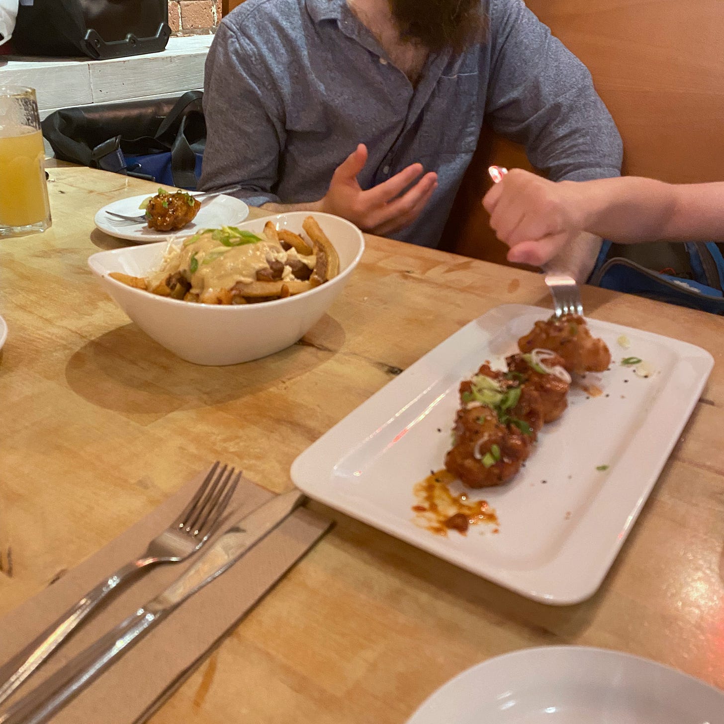 A rectangular plate of sweet chili cauliflower, dusted with green onions and sesame. Tyrion's hand sticks a fork into one piece. Next to it is an oblong bowl of poutine, and Jeff sits behind it, talking to Tyrion whose face is out of frame. In the foreground is a napkin set with a knife and fork, and the corner of a small side plate is visible.