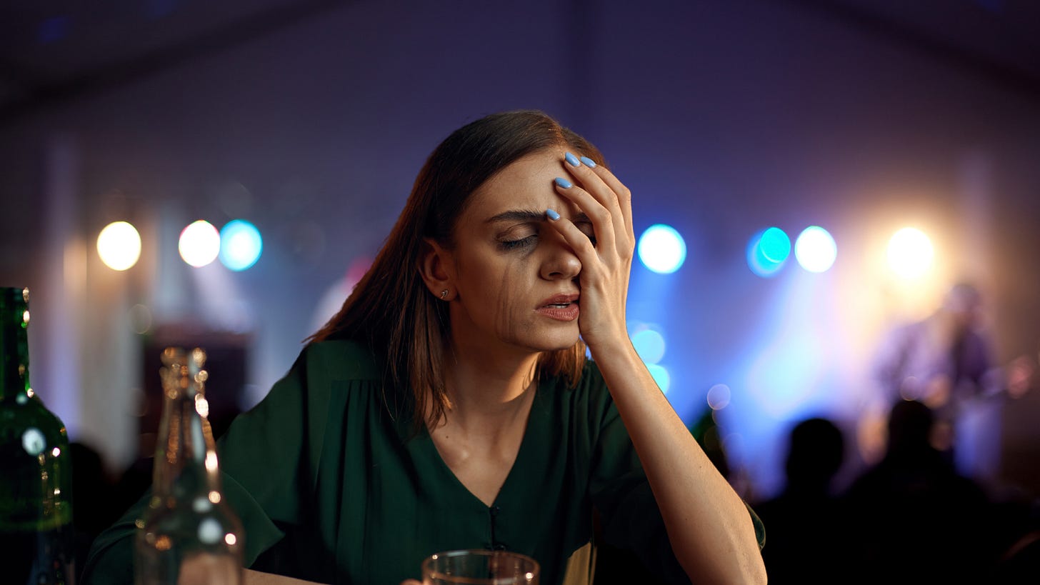 A distressed woman with tear-streaked makeup sits at a table, her hand covering her face in emotional exhaustion. In the blurred background, a band plays under soft, glowing lights. This image symbolizes Pisces' emotional depth, escapism, and the struggle with overwhelming feelings