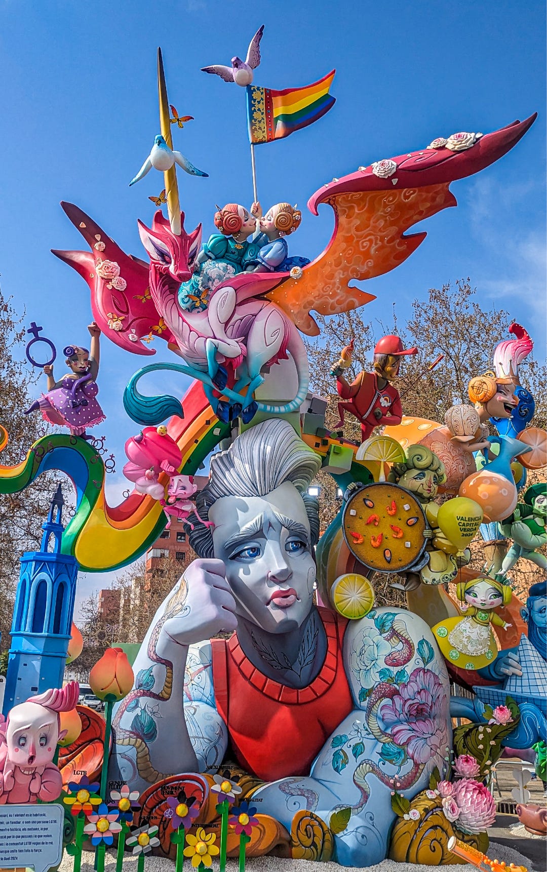 Closeup of Mama, soy gay focusing on pensive young man surrounded by smaller figures, rainbow flags, and overhead a pink and orange unicorn with two lesbians kissing