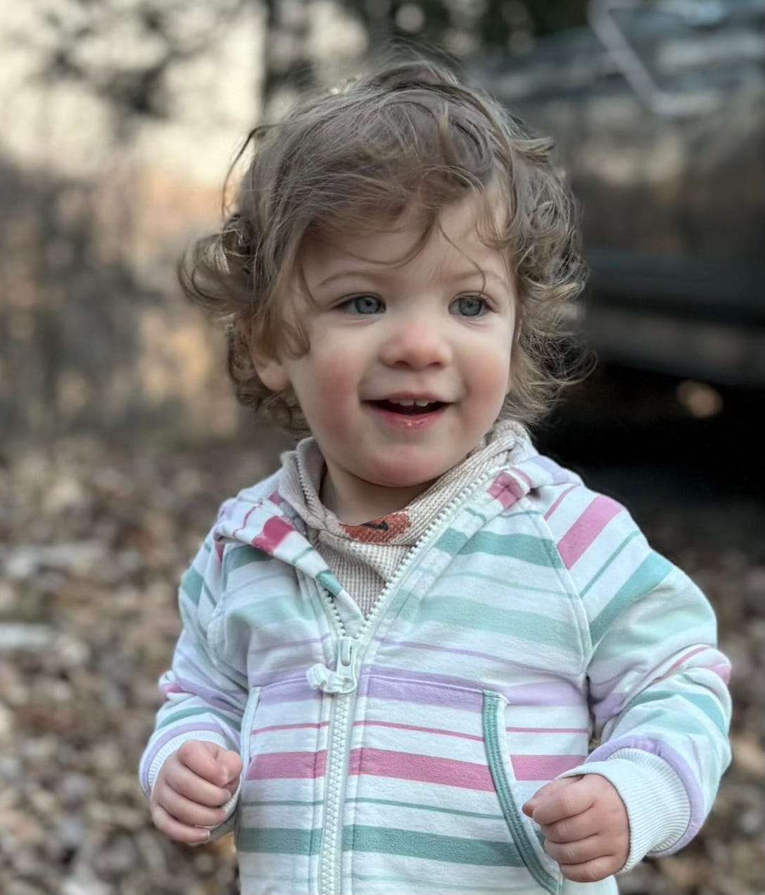 A little girl with curly brown hair and a pastel striped hoodie stands in a tree-lined gravel driveway, smiling.