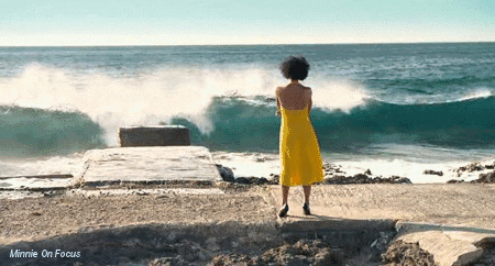 gif animado de vídeo em espaço aberto. mar revolto numa praia em que uma mulher negra de vestido amarelo observa as ondas quebrando contra a beira.