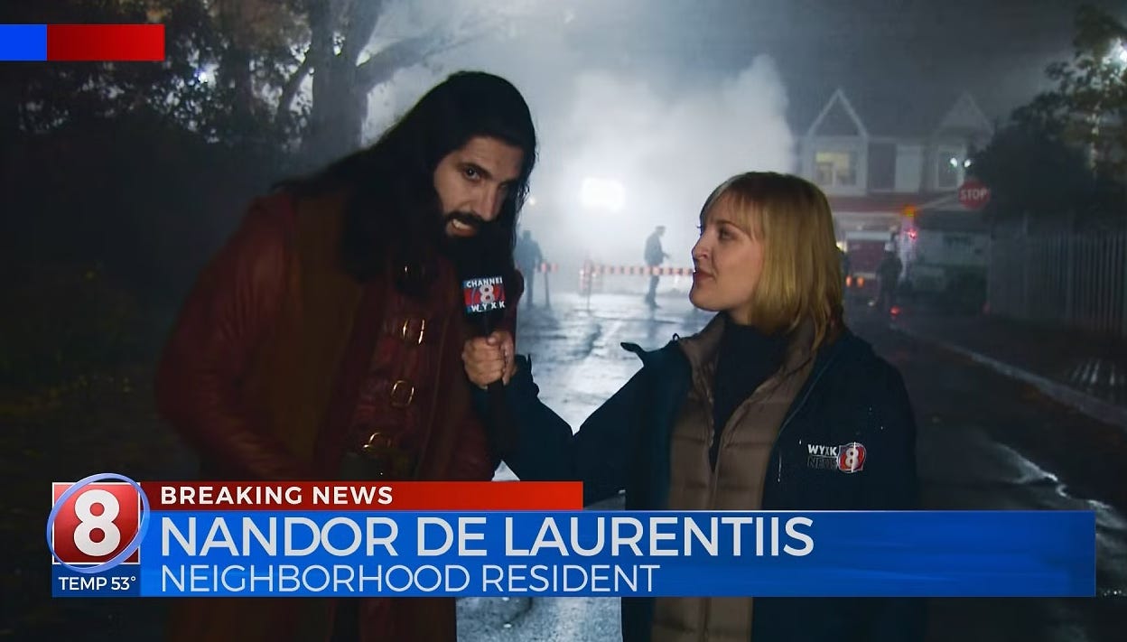 Nandor speaks to a local news anchor on a residential street; a chyron reads "Breaking News, Nandor De Laurentiis, Neighborhood resident" a la a local news segment 