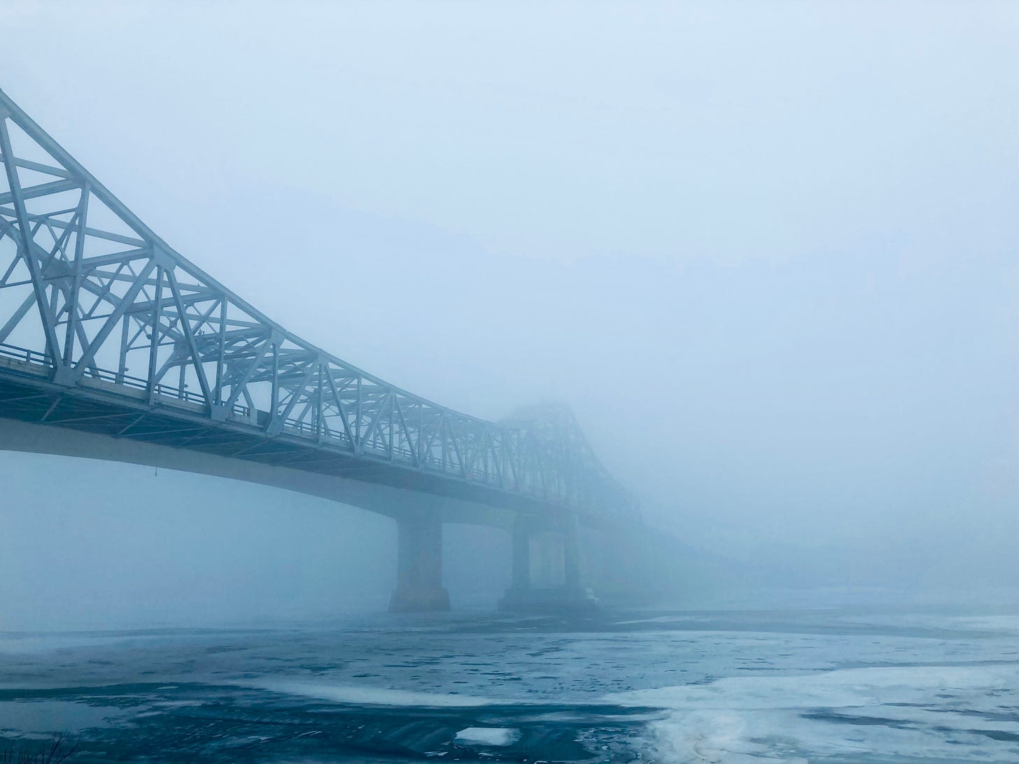 An iron bridge spans an icy river, stretching into a dense fog