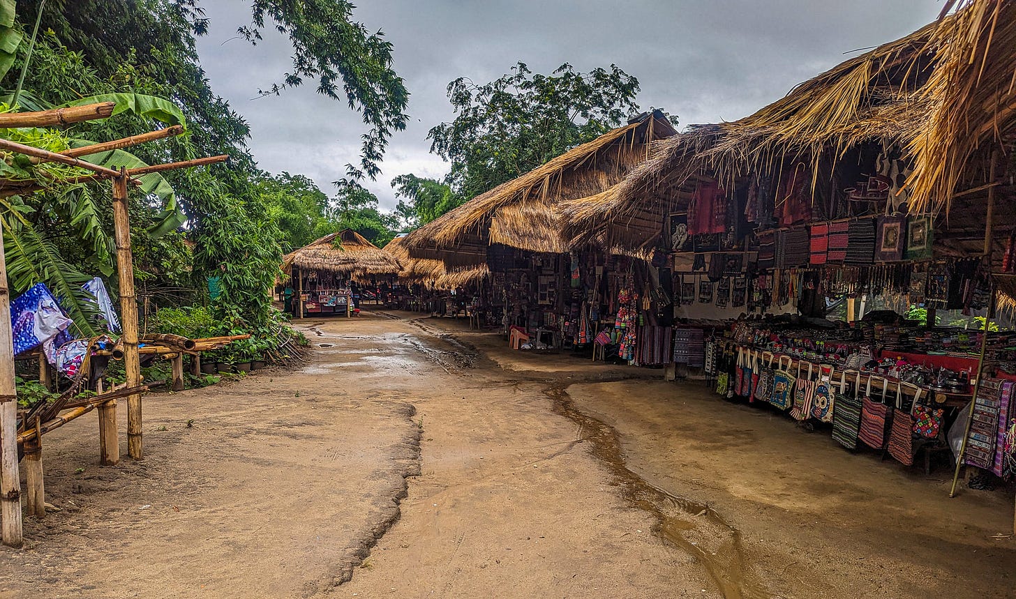 Thatch-roofed stalls filled with hand woven bags and clothing overlooking a muddy dirt path. 
