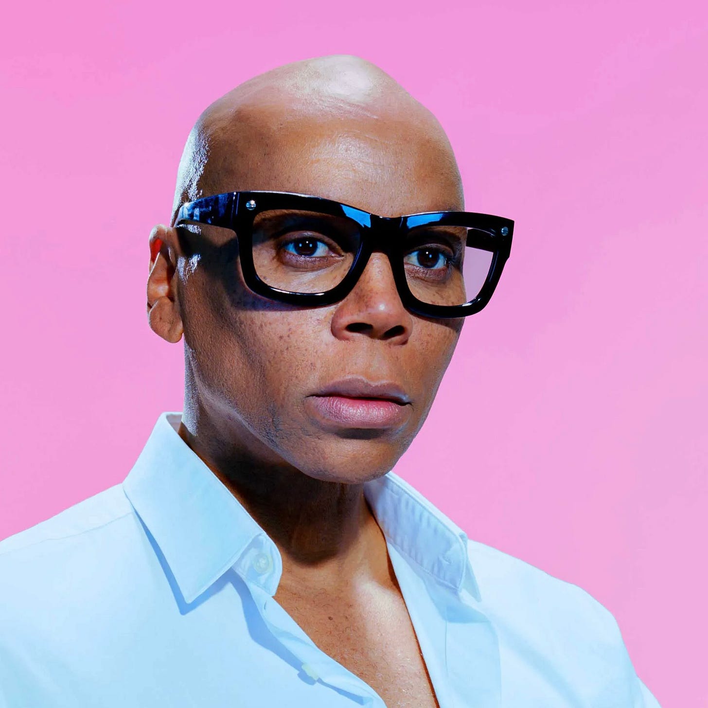 Photo of RuPaul in thick black glasses and a light blue shirt, in front of a pink background