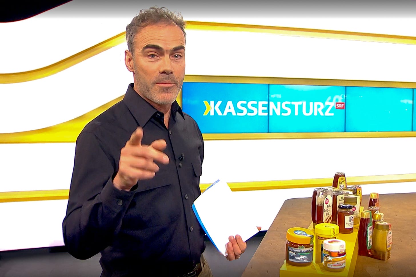 Das Bild zeigt den Moderator André Ruch in der Schweizer TV-Sendung «Kassensturz» auf SRF. Er steht im Studio und hält ein Notizheft in der Hand, während er in die Kamera spricht. Im Hintergrund ist das «Kassensturz»-Logo auf einem großen Bildschirm zu sehen. Auf dem Tisch vor ihm stehen mehrere Honiggläser und Honigspender. Die Szene ist Teil einer Anmoderation zu einer Reportage über Importhonige, die mit Zuckersirup verfälscht wurden. Das Bild zeigt den Moderator André Ruch in der Schweizer TV-Sendung «Kassensturz» auf SRF. Er steht im Studio und hält ein Notizheft in der Hand, während er in die Kamera spricht. Im Hintergrund ist das «Kassensturz»-Logo auf einem großen Bildschirm zu sehen. Auf dem Tisch vor ihm stehen mehrere Honiggläser und Honigspender. Die Szene ist Teil einer Anmoderation zu einer Reportage über Importhonige, die mit Zuckersirup verfälscht wurden.