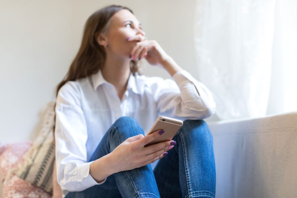 Young woman looking out a window contemplatively while holding her cell phone.