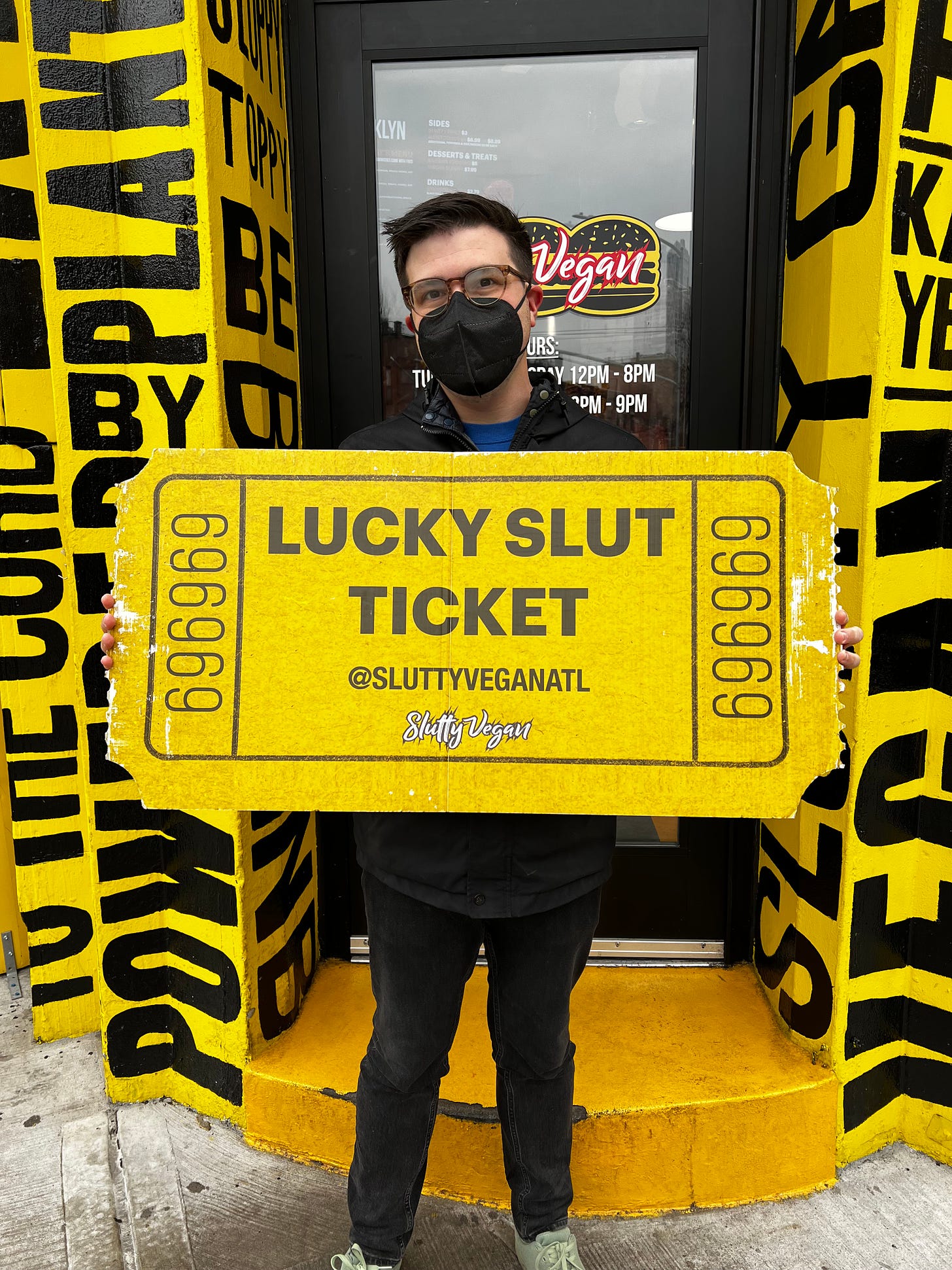 The author holding large yellow "Lucky Slut Ticket" in front of Slutty Vegan.