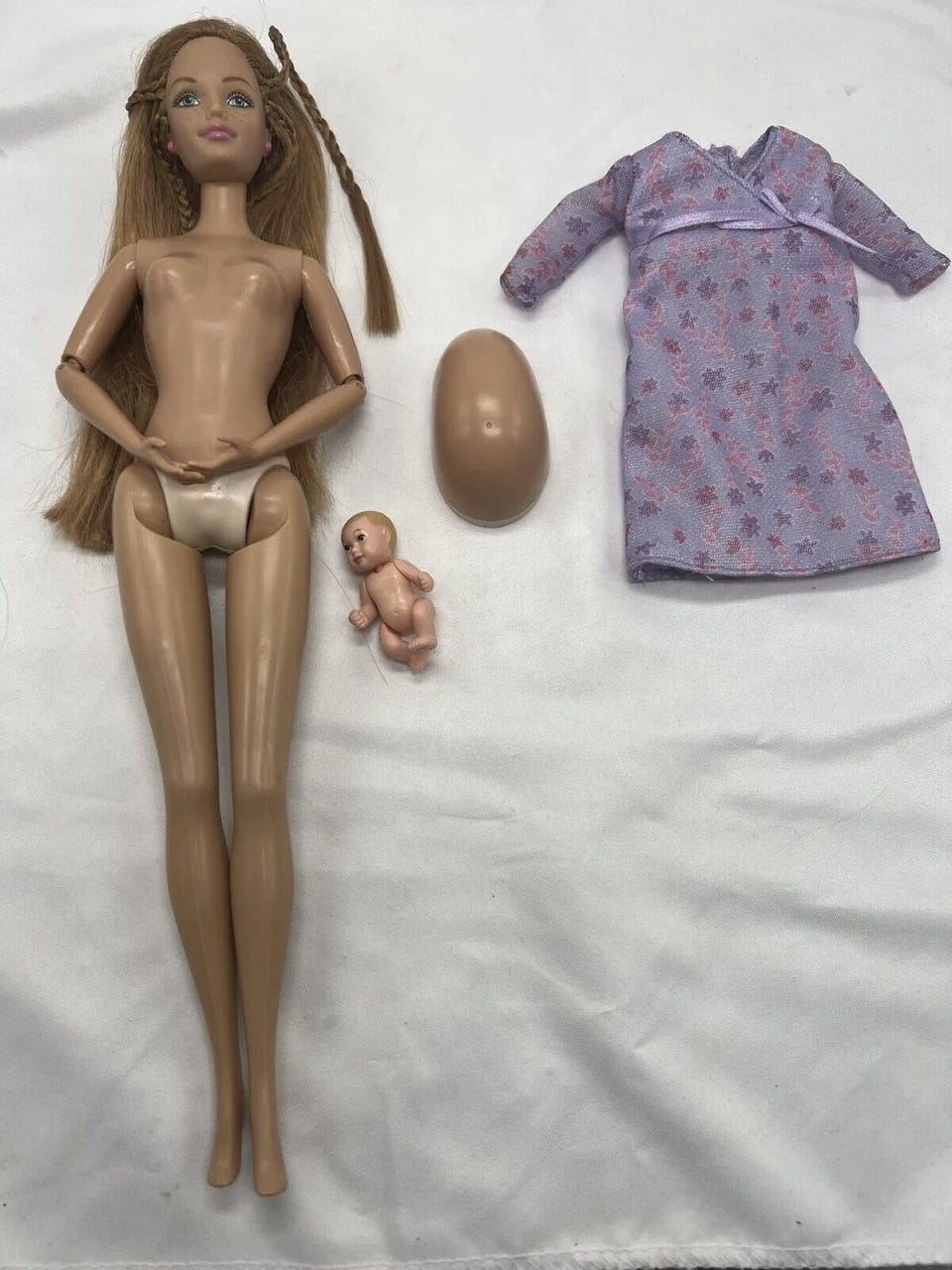 A flat lay of a naked Barbie doll, a removable pregnant stomach, a small baby, and a dress.