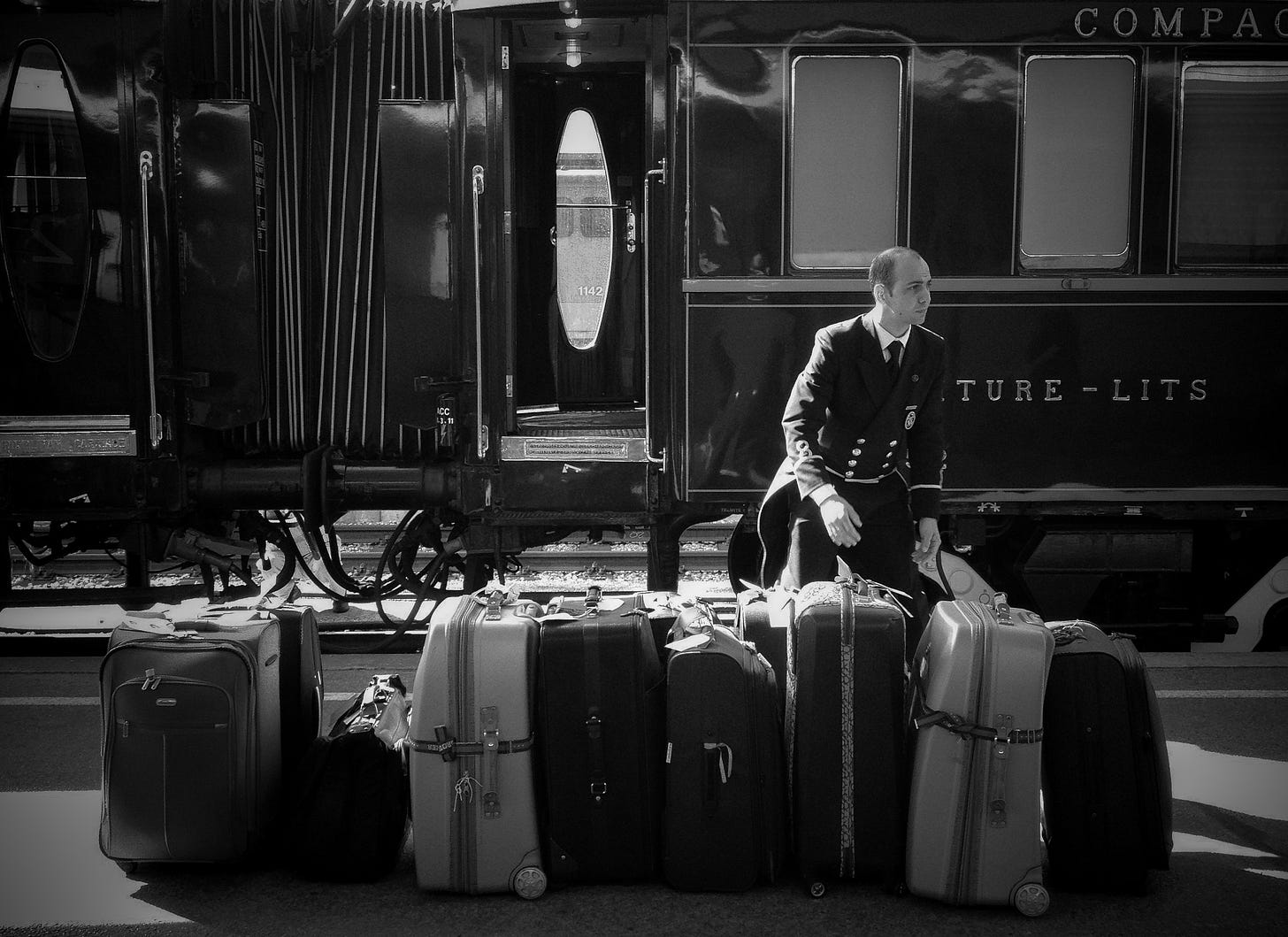 A smartly dressed baggage handler wrangles a row of suitcases stood on the platform next to the stationary carriages of the Orient Express.