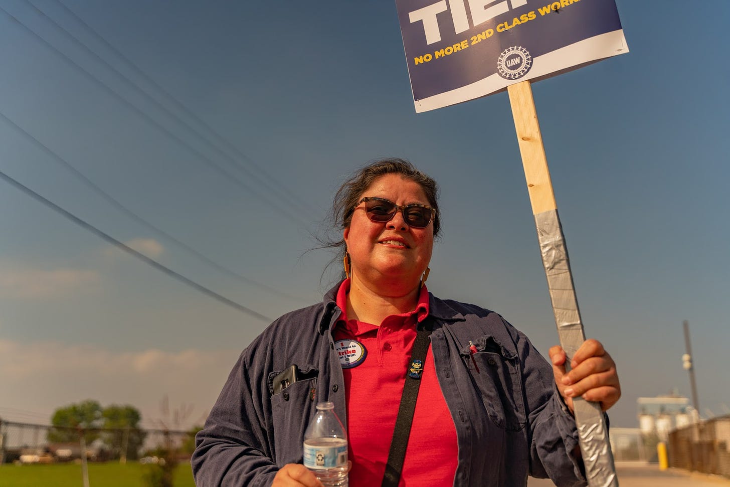the upper half of a smiling woman with dark brown hair wearing sunglasses, a red polo shirt and dark blue outer shirt, holding a picket sign and a plastic water bottle