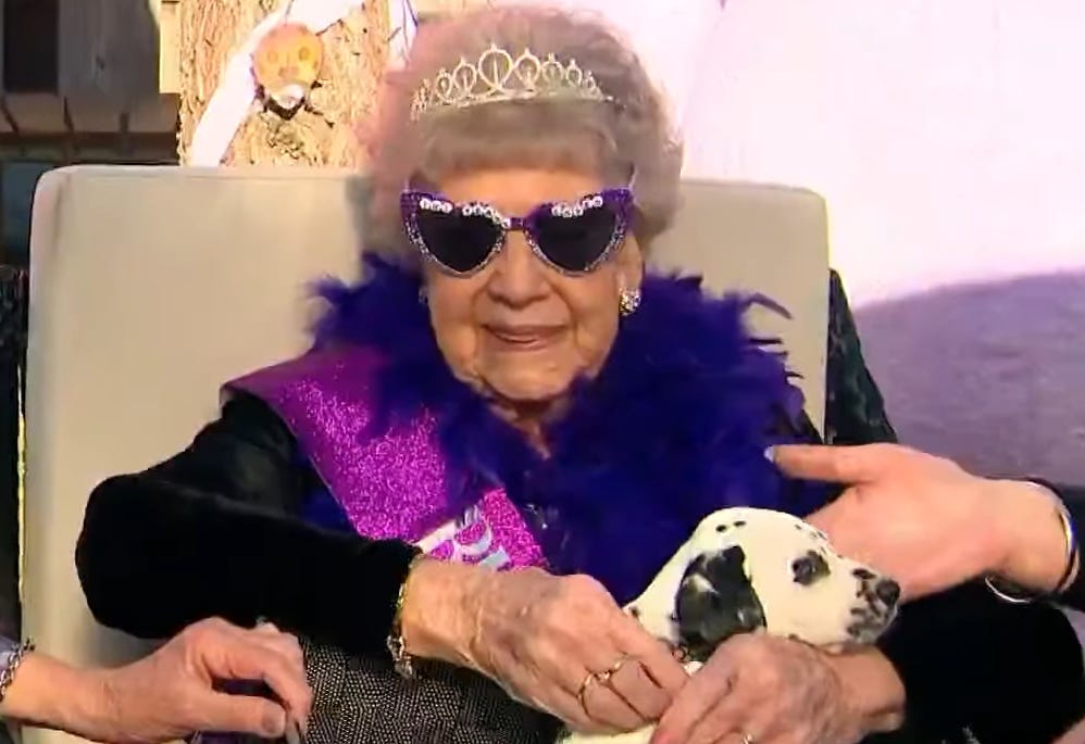 An old lady sat on a chair with large purple sunglasses on, a tiara on her head and a Dalmation puppy on her lap. She is smiling.