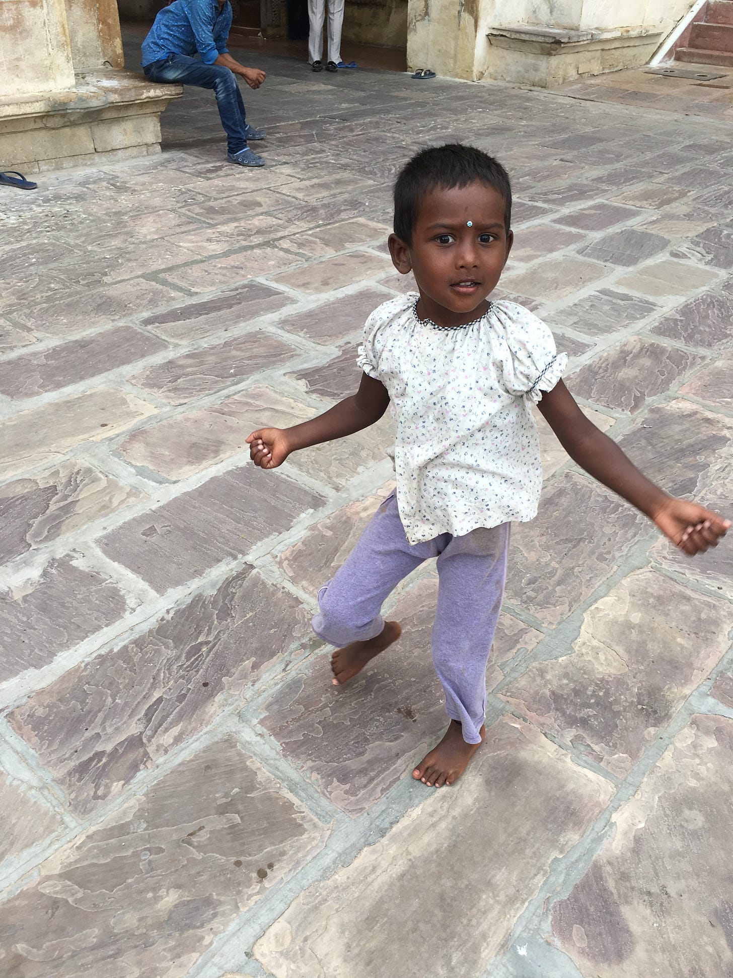 A three-year-old girl twirls in a stone and marble courtyard. She has very short black hair, dark brown skin, and dark eyes. She wears a white shirt and purple pants, looking off into the distance.