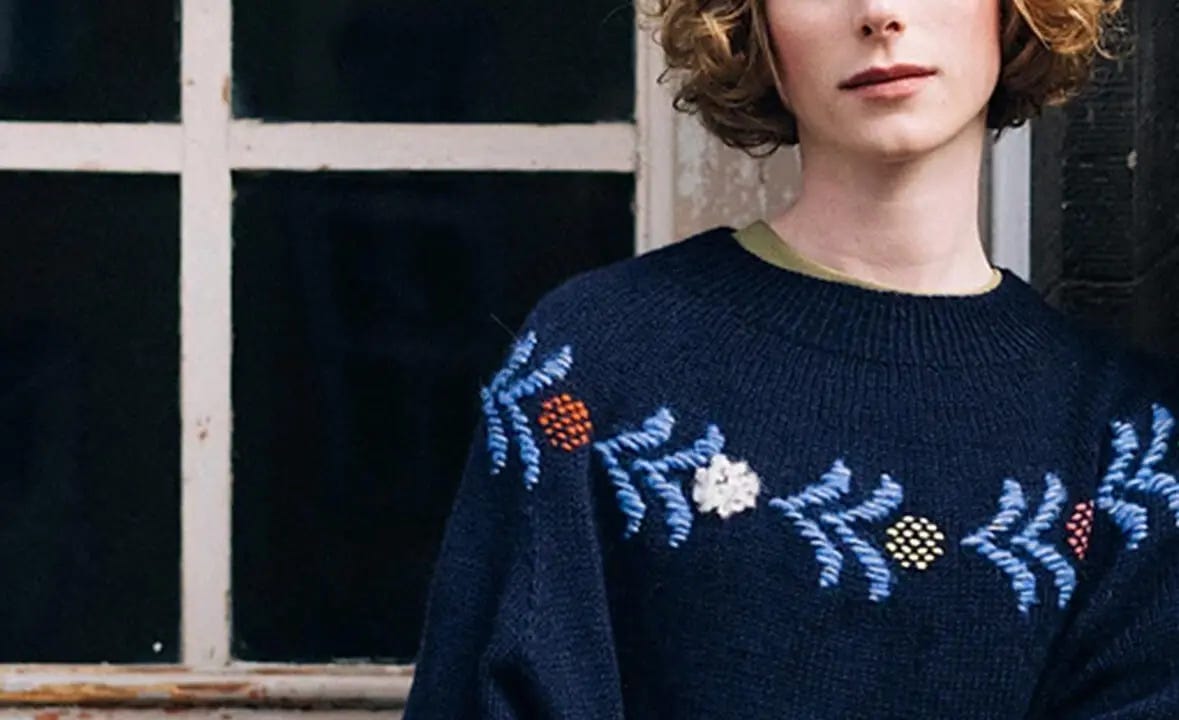 woman stands in front of window wearing a navy blue sweater with light blue, white, yellow and orange roosimine designs