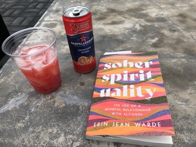Erin's book Sober Spirituality sits on a stone table beside a San Pellegrino Blood Orange Soda can, with a cup of ice and blood orange soda