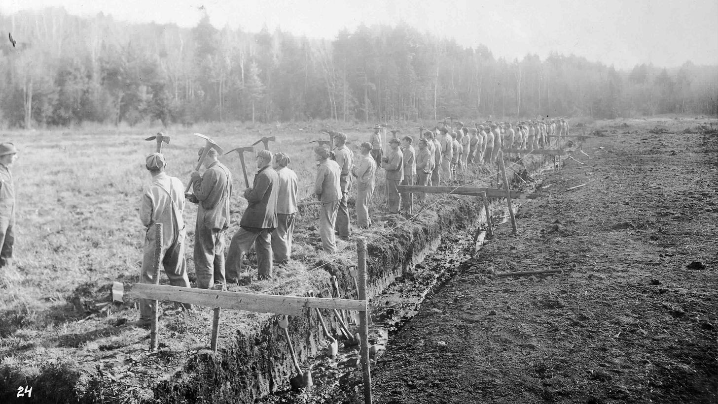 Black and white photograph of young men in a row, backs turned, with pickaxes, standing next to a ditch in a large field.