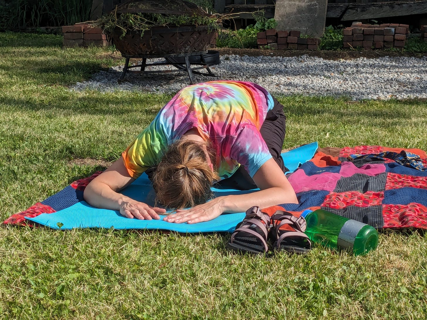 A woman in a tie dye shirt does a child's pose on a yoga mat in a back yard. Her mat is on a quilt, and there are sandals and a water bottle directly in front of the mat.