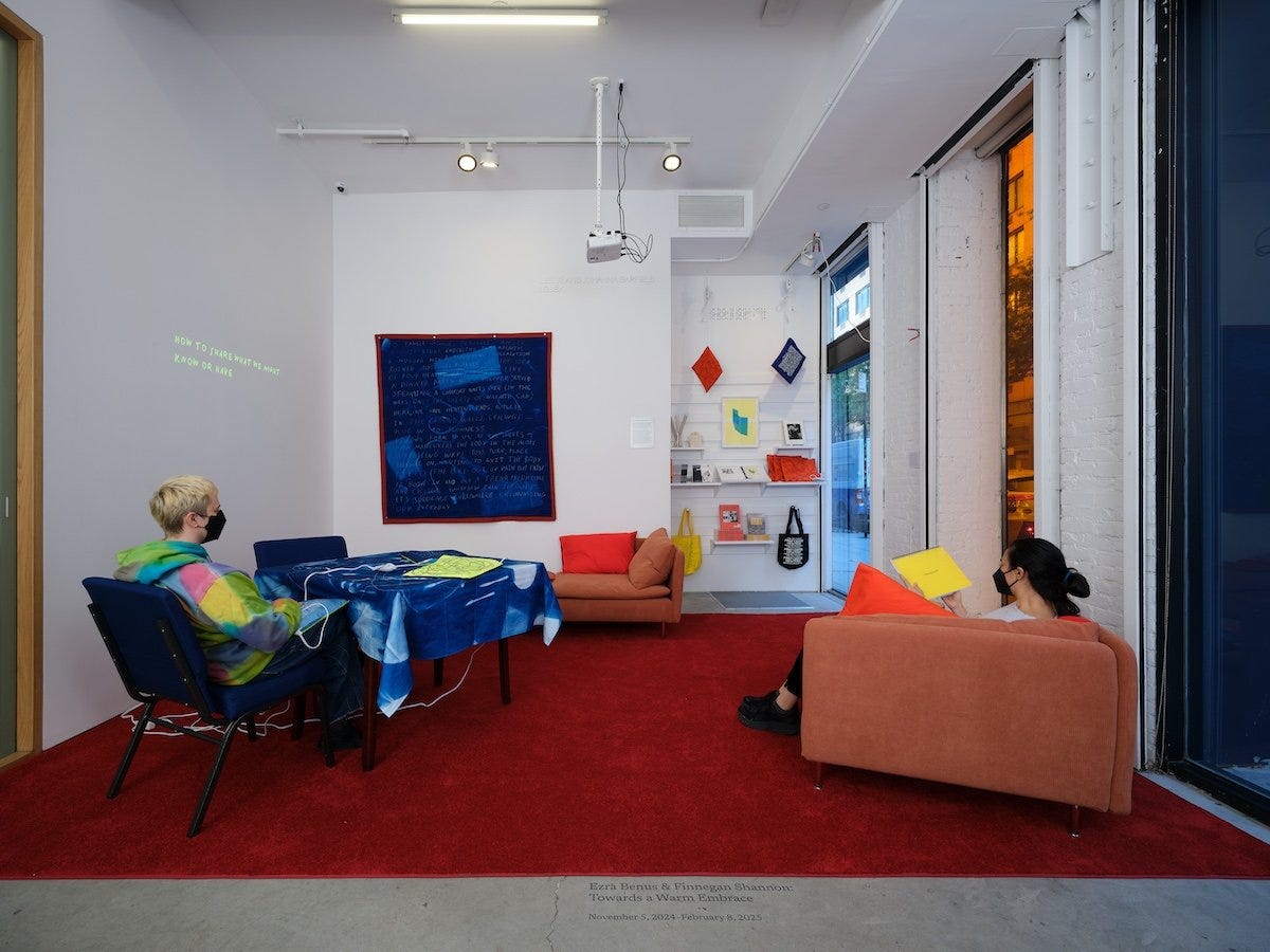 A room with a cozy setup including two orange couches and a table covered in a blue cyanotype cloth and heating pads on top. In the background, colorful prints and textiles hang on the wall, while visitors sit on the couches.