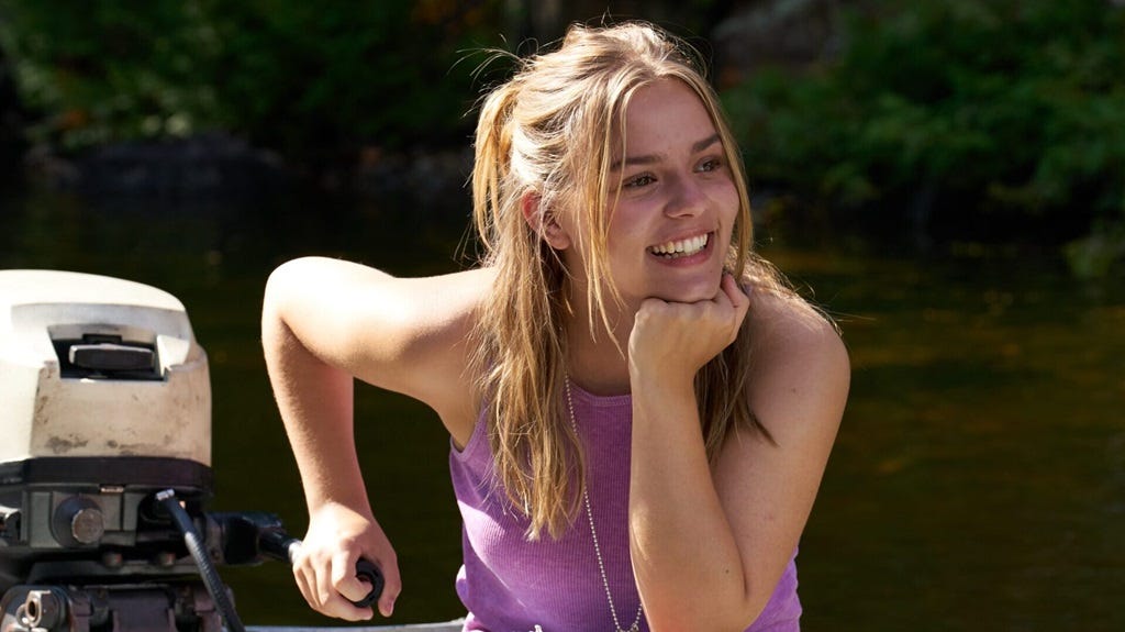 A woman is shown driving a boat. One hand is on the outboard motor. She smiles.