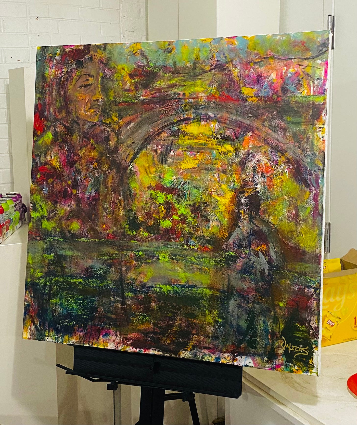 A painting by Onicas Gaddis with lots of color and texture displayed on a black metal easel. The painting features a large face in the top left corner, a bridge, and a person with their back to the viewer in the bottom right corner.