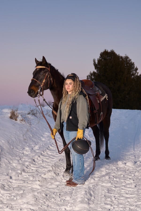 A woman with her hair styled in locs standing next to and holding the reins of a dark bay horse on a snow-covered landscape