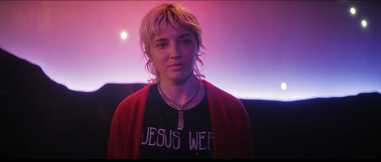 A person stands center frame. They have short, blond hair, and wear a red cardigan, a black t-shirt with the words “Jesus Wer” showing, and a chain link necklace. Behind them is a background featuring a cut-out of a canyon underneath a gradient of pink and purple, with lights resembling stars.