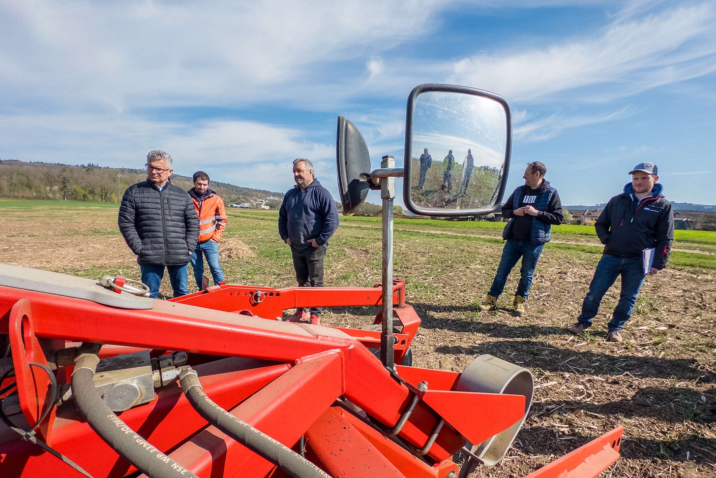 Teilnehmer eines Bodenkurses für regenerative Landwirtschaft stehen auf einem Acker vor einer roten Bodenfräse, die im Vordergrund zu sehen ist. In der Bildmitte befindet sich Kurs-Organisator Sven Studer. Im Rückspiegel der Maschine spiegeln sich weitere Teilnehmer der Veranstaltung. Teilnehmer eines Bodenkurses für regenerative Landwirtschaft stehen auf einem Acker vor einer roten Bodenfräse, die im Vordergrund zu sehen ist. In der Bildmitte befindet sich Kurs-Organisator Sven Studer. Im Rückspiegel der Maschine spiegeln sich weitere Teilnehmer der Veranstaltung.