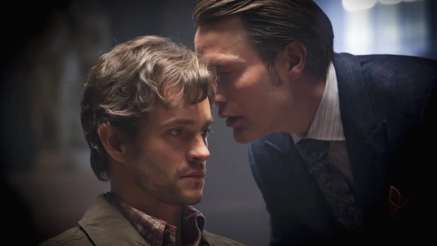 A man played by Mads Mikkelsen, wearing coiffed hair and a dark blue suit, whispers in the ear of a man played by Hugh Dancy, wearing a flannel shirt and jacket