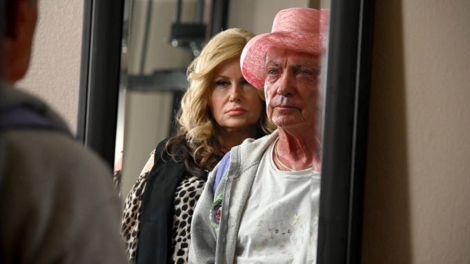 The viewer looks into a tall mirror with an older white man wearing a ratty white tee-shirt under a grey zip-up sweatshirt with a fancy pink old-ladies-hat on his head. Looking over his shoulder, also into the mirror, is a blonde 40-something white woman who is regarding him with A LOOK. He looks back at her gaze in the mirror with a firm gaze and a neutral expression.
