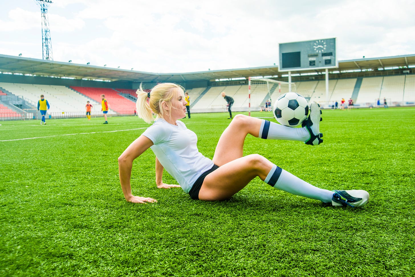 Woman in white T-shirt on ground balancing soccer ball on her foot.