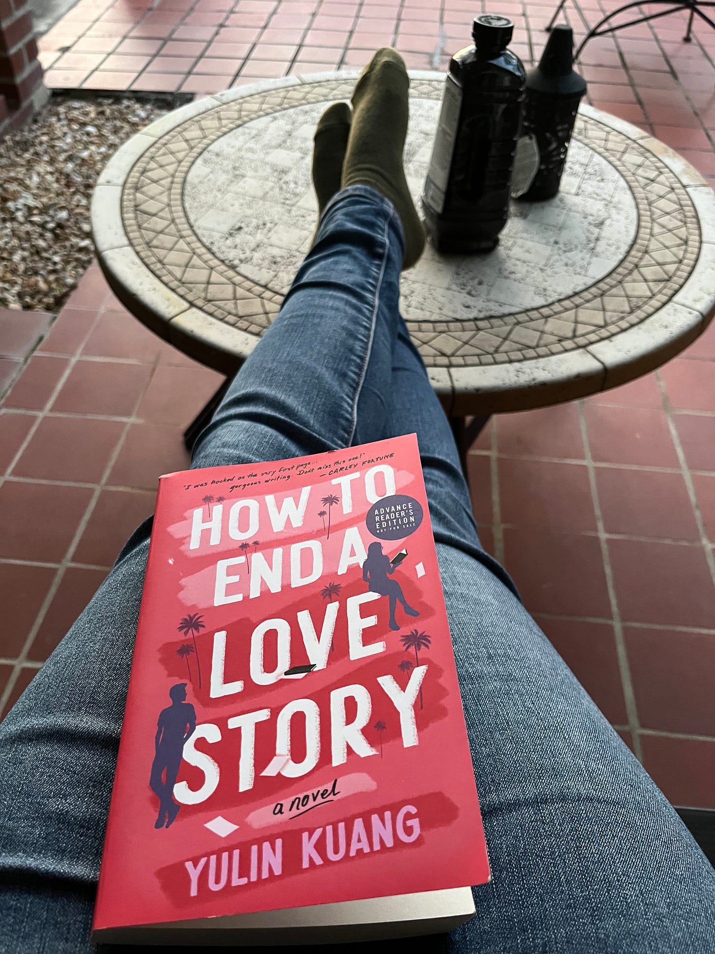 Picture of my legs stretched out and resting on a patio table, green socks on my feet and jeans covering my legs, with an advance copy of HOW TO END A LOVE STORY in my lap
