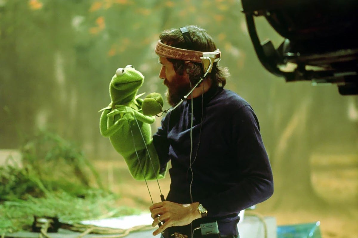 A bearded white man with dark hair holds up a Kermit puppet, while a camera films him.