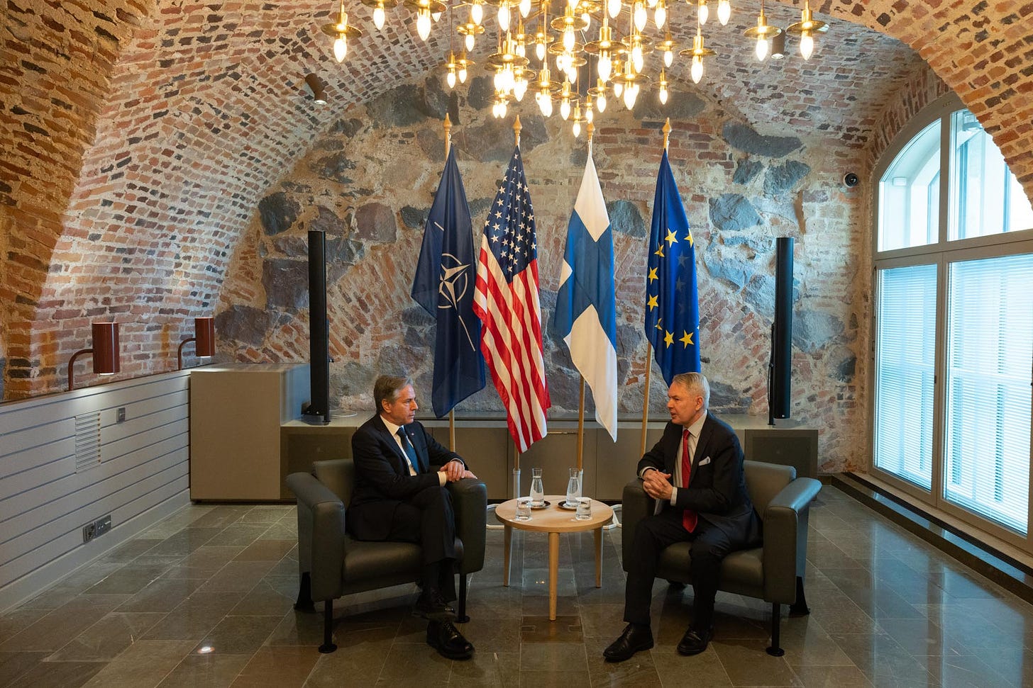 Secretary Blinken and Foreign Minister Haavisto sit in upholstered chairs with a small wooden table in the middle. Four flags sit behind them.