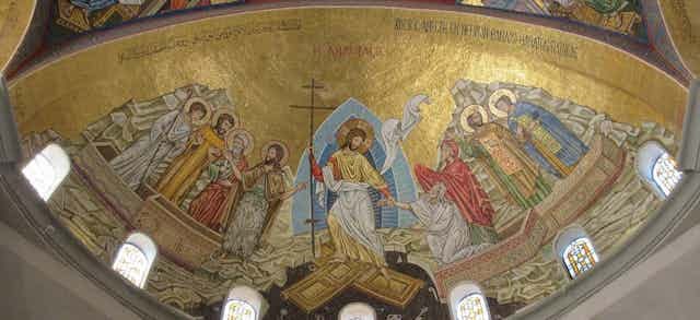 A mosaic with a golden background, six figures with halos, Jesus holding a cross in the middle, and two people whom he is helping out of coffins.