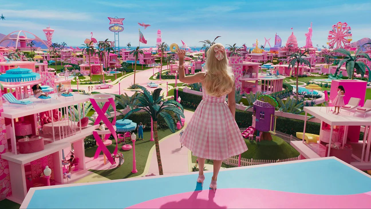 Movie still from Barbie. Barbie stands on the roof of her home and waves at Barbieland laid out before her.