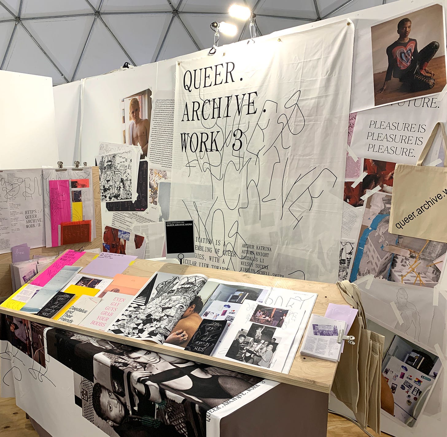 A view of the Queer.Archive.Work booth at the New York Art Book Fair in 2019. In the foreground is a slanted wood display with many pamphlets and zines layered over one another. In the background is a wall with many more publications, images, and totes in a jumble. A large cloth sign or poster says QUEER.ARCHIVE.WORK in both typed and hand-drawn letters. The effect is exuberantly varied and productive.