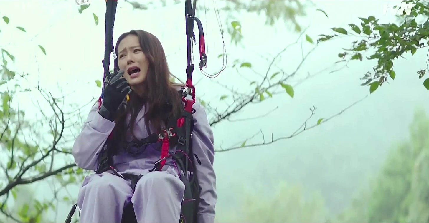 A Korean woman hangs from her paragliding equipment. She is stuck in a tree. She is visibly upset as she attempts to contact someone on her walkie-talkie.