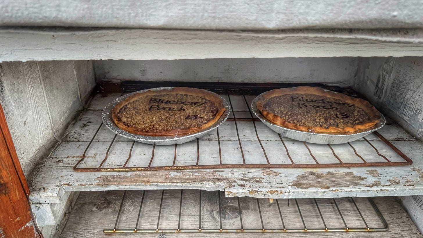 Closer shot of the interior of one of the stand's compartments. There are two blueberry-peach pies in aluminum tins and wrapped with foil sitting on a metal oven rack inside a whitewashed but worn compartment. 