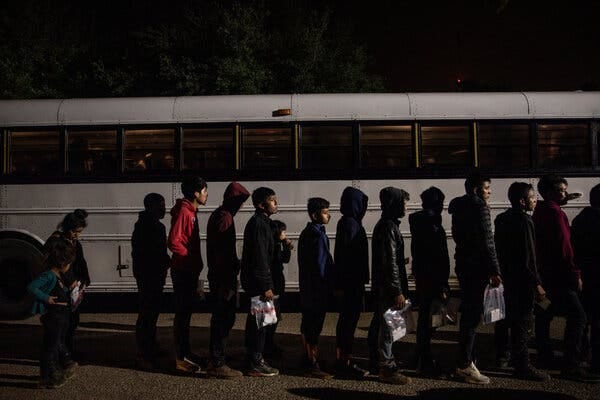 A line of unaccompanied migrant children waiting to board a white bus.