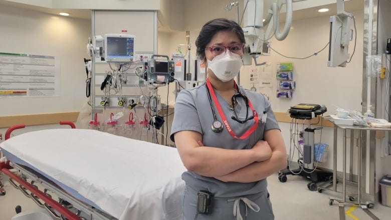Dr. Katie Lin is dressed in grey scrubs and a face mask as she stands in front of an ER treatment bed with her arms crossed.