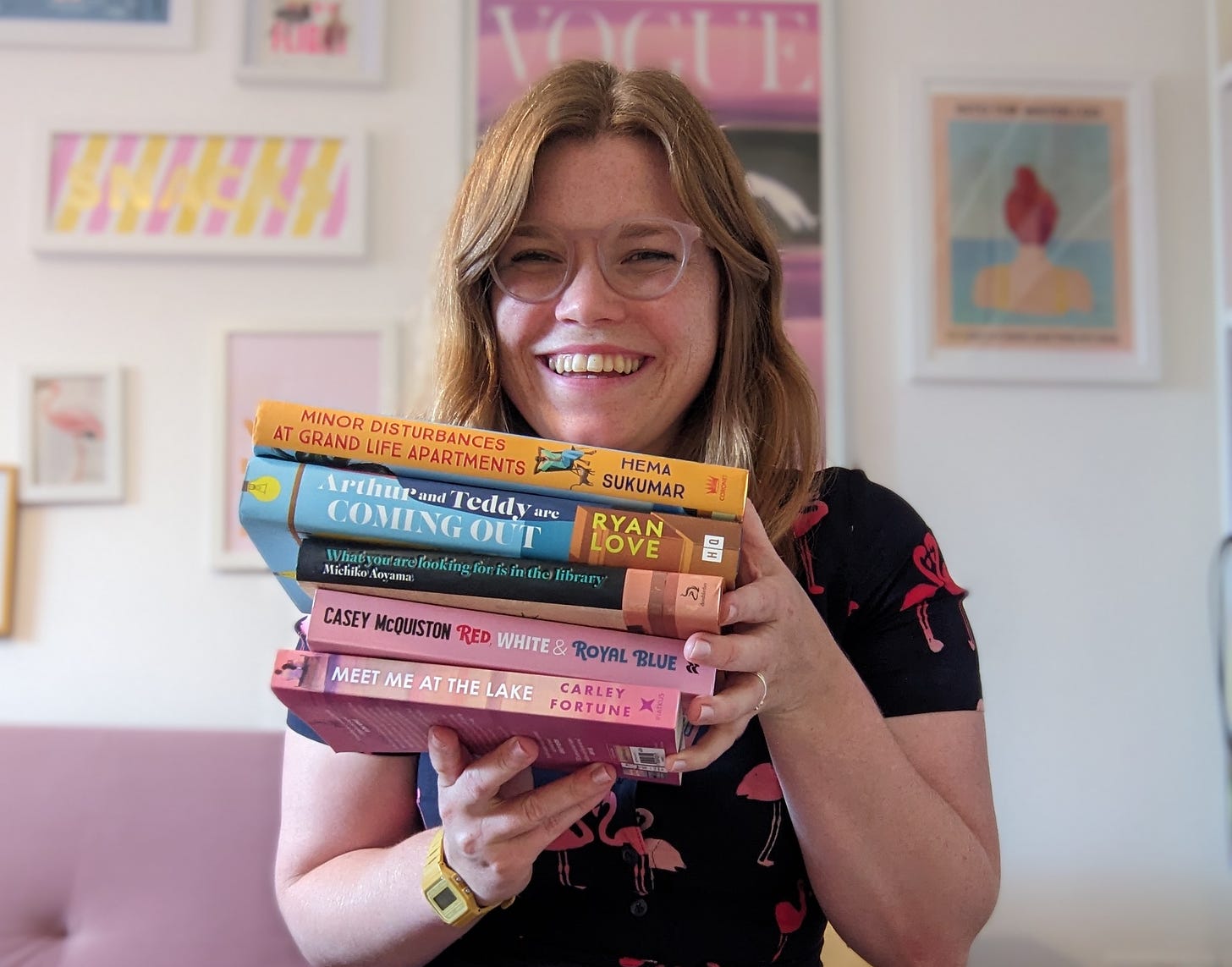 Libby Page wearing a flamingo-print dress and holding a stack of books. She is smiling and wearing glasses. 