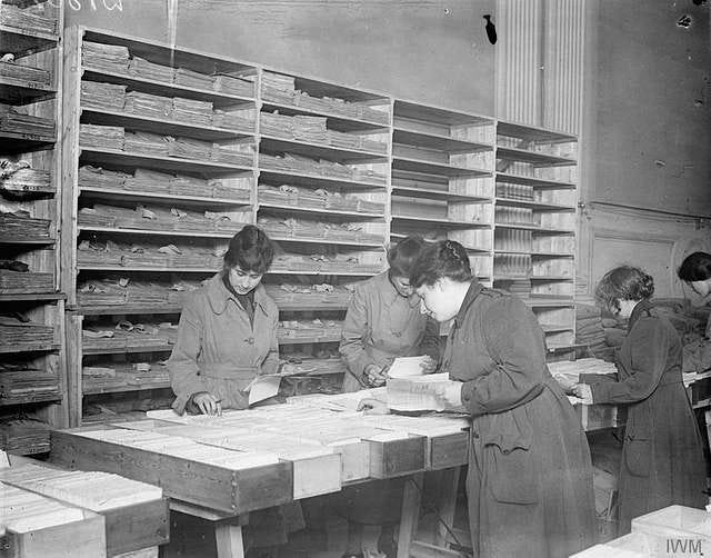 Black and white photo of four women engaged in some serious looking paperwork. They appear to be looking intently at papers in their hands and filing them into wooden boxes of record cards on a trestle table. Behind them shelves of record boxes cover the wall.