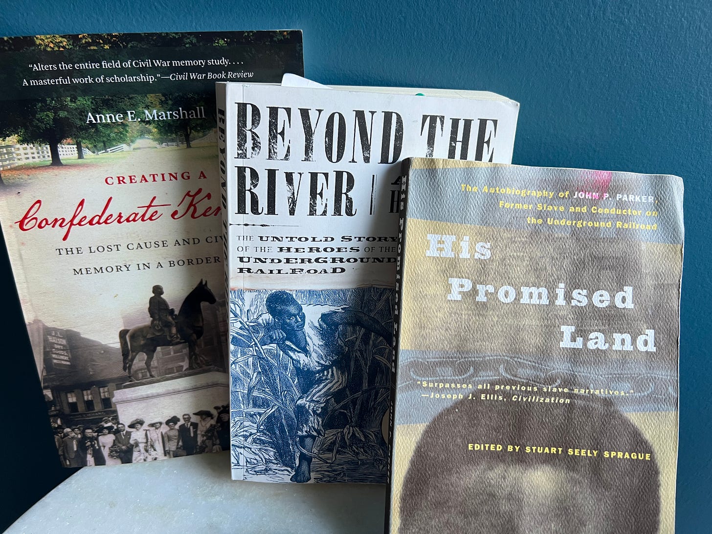 Three books against a blue background. "Creating a Confederate Kentucky," "Beyond the River," and "His Promised Land"