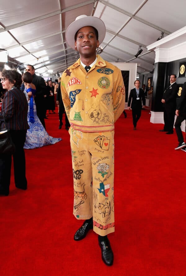 Leon Bridges, standing on a red carpet, wearing a white Western hat, a yellow corduroy suit decorated with colorful hand-drawn illustrations, a collared shirt and black shoes. 