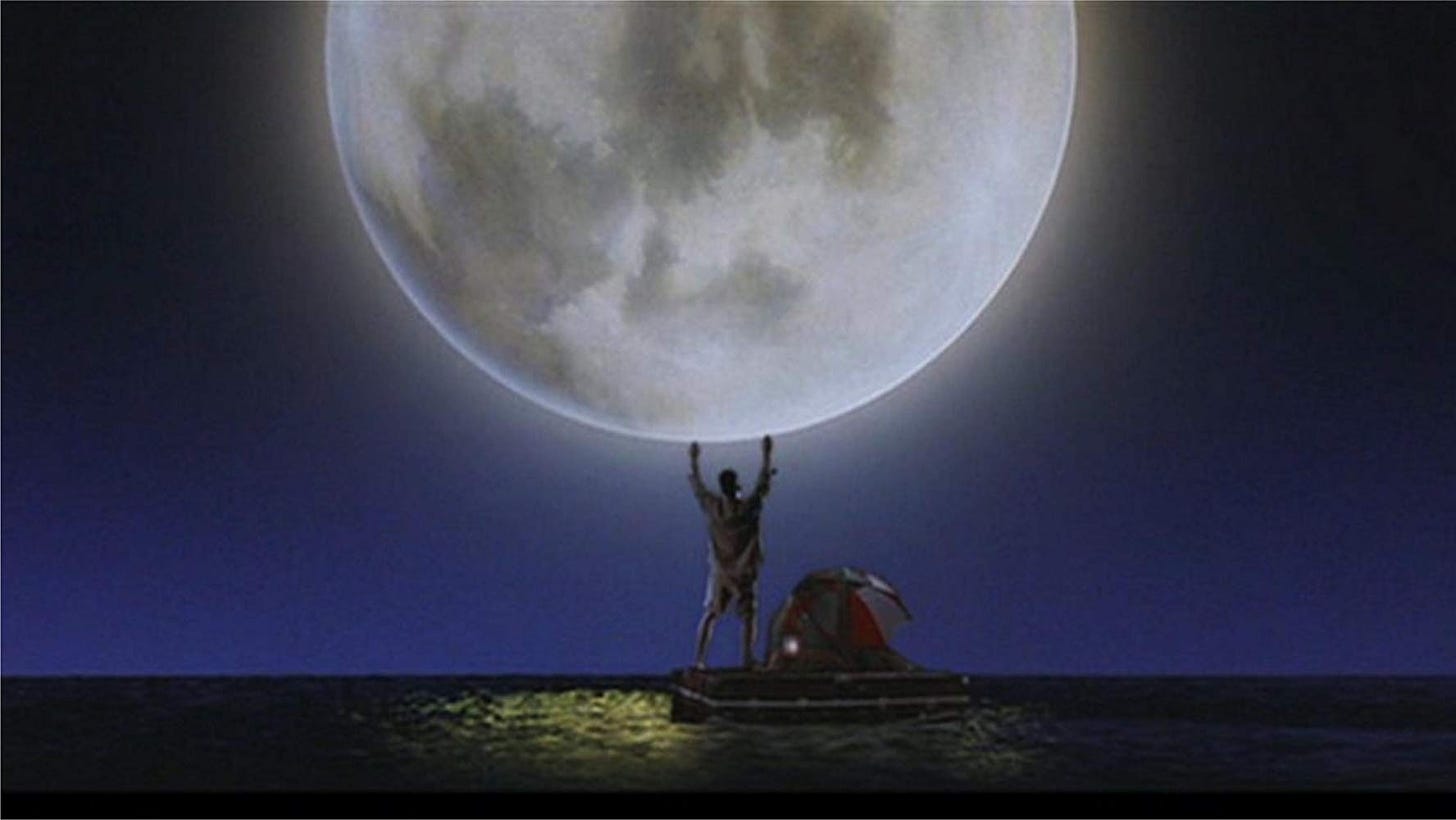 Still frame from the movie, “Joe vs the Volcano”. Tom Hanks stands alone atop his steamers trucks, arms high as the full moon raises on the ocean’s horizon