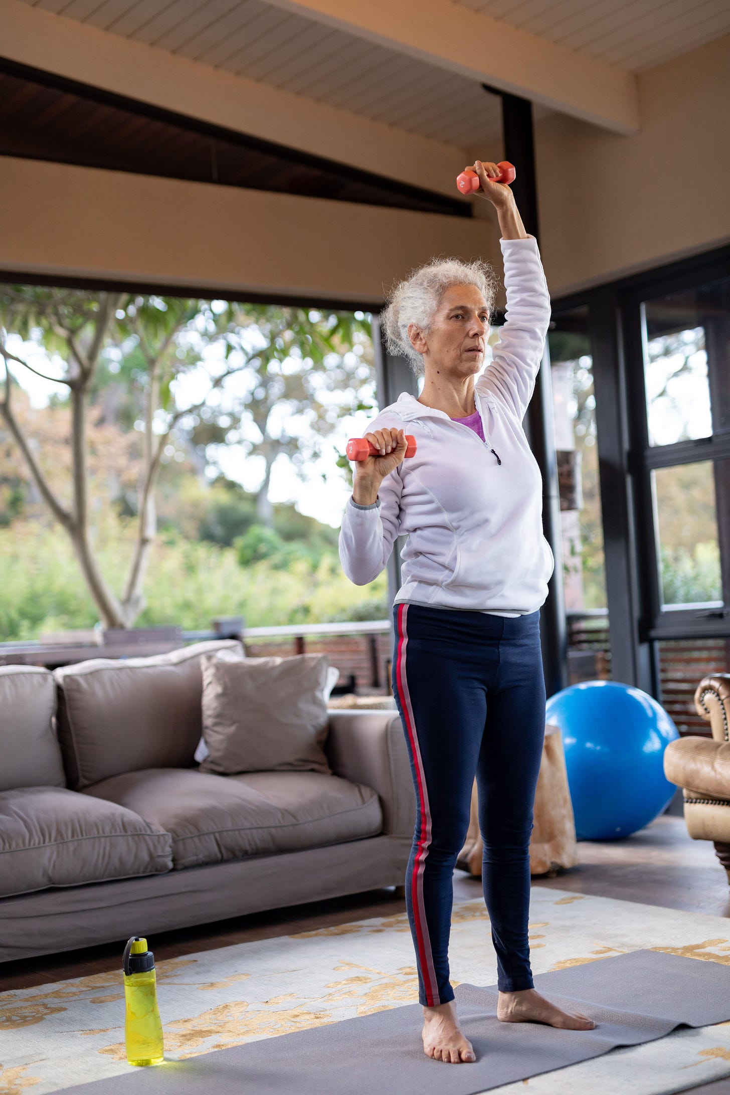 Senior caucasian woman in living room exercising, standing and lifting dumbbells