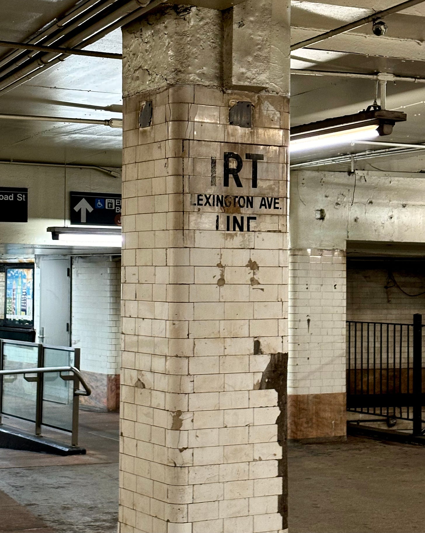 A white tiled column in a subway station. In black letters it says IRT LEXINGTON AVE. LINE in slightly battered lettering.