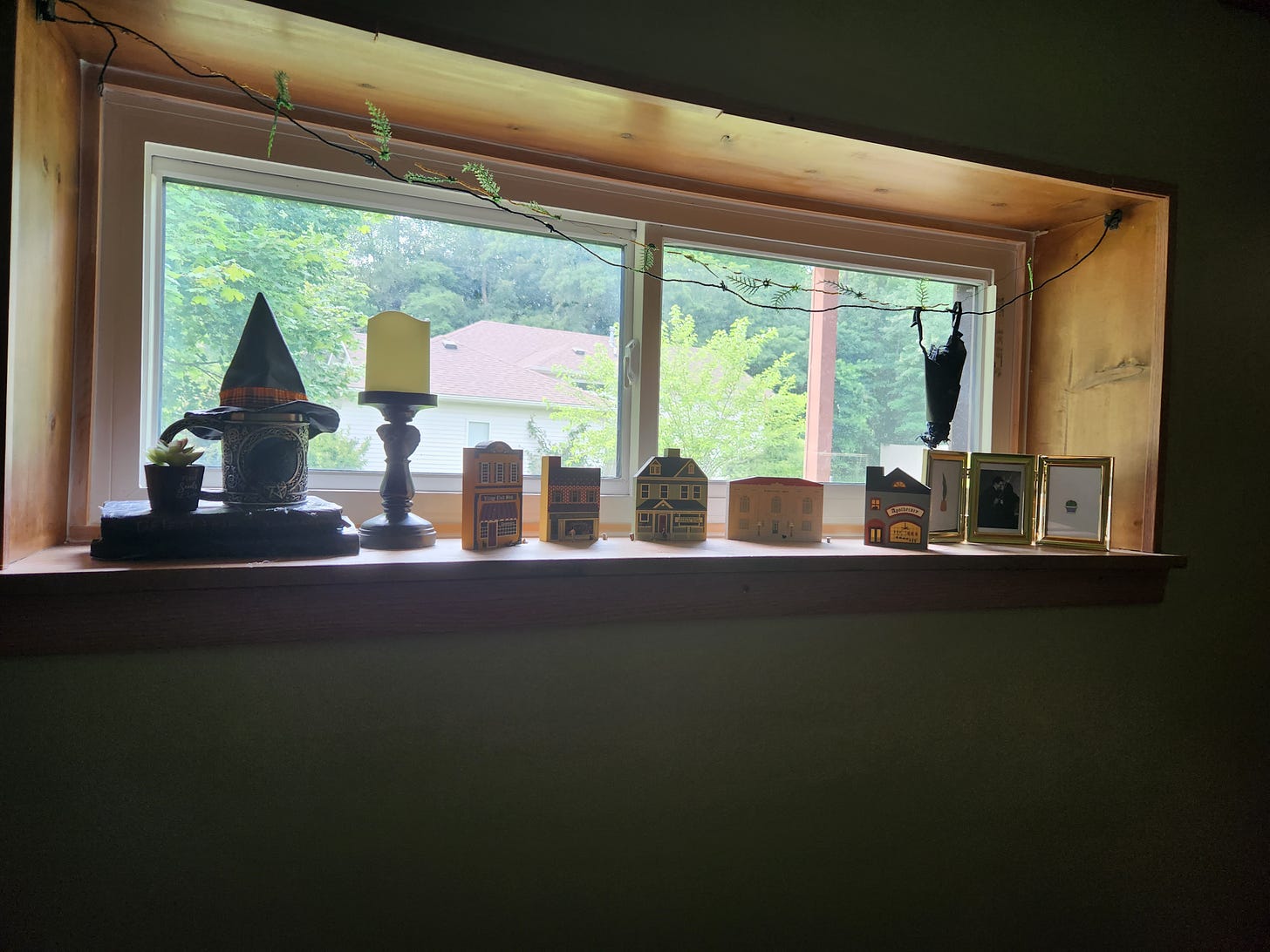 A huanted window display in Kandi's kitchen. A hanging bat, a spooky mug, a haunted village, old spell books, a witch's hat, a candle holder in the shape of a crow, and a picture frame with Gomez and Morticia are all present.