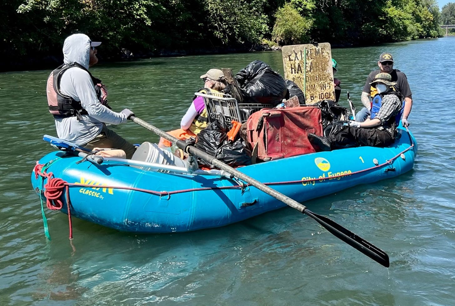 Three people on a blue raft that is full of trash, including a grocery cart, suitcase, buckets, and an old metal sign.