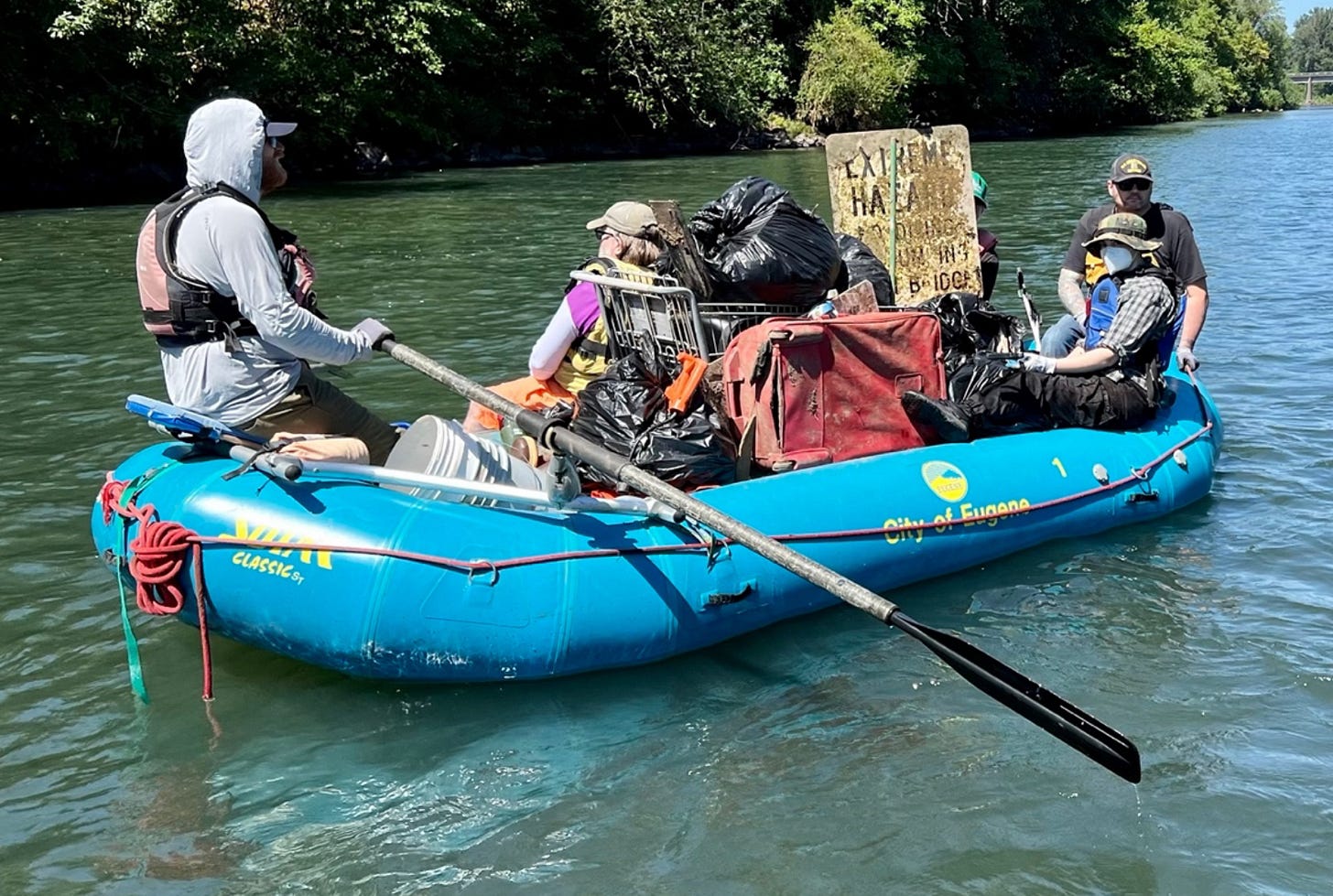 Three people on a blue raft that is full of trash, including a grocery cart, suitcase, buckets, and an old metal sign.